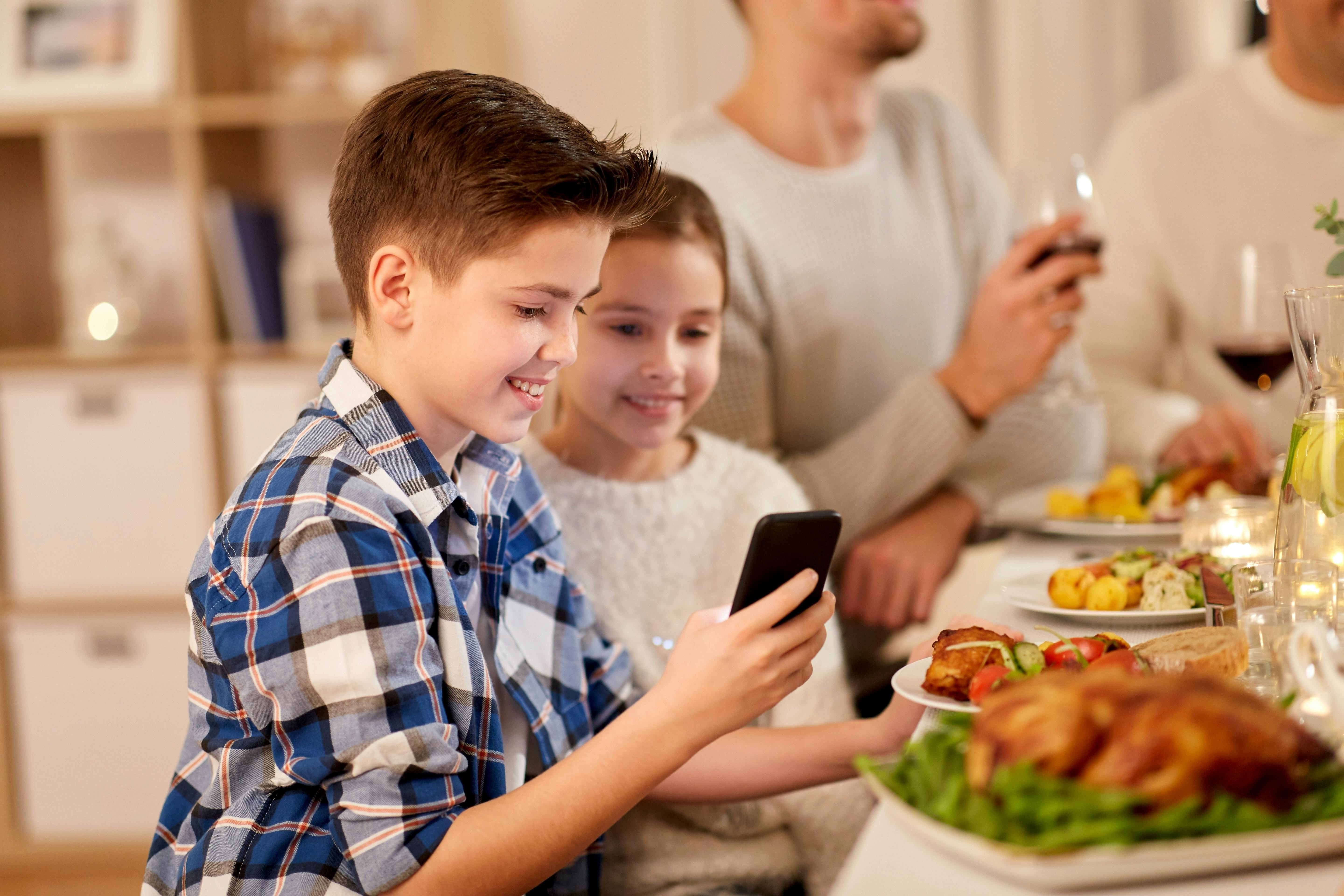 technology, holidays and people concept - happy boy with sister using smartphone having family dinner party at home