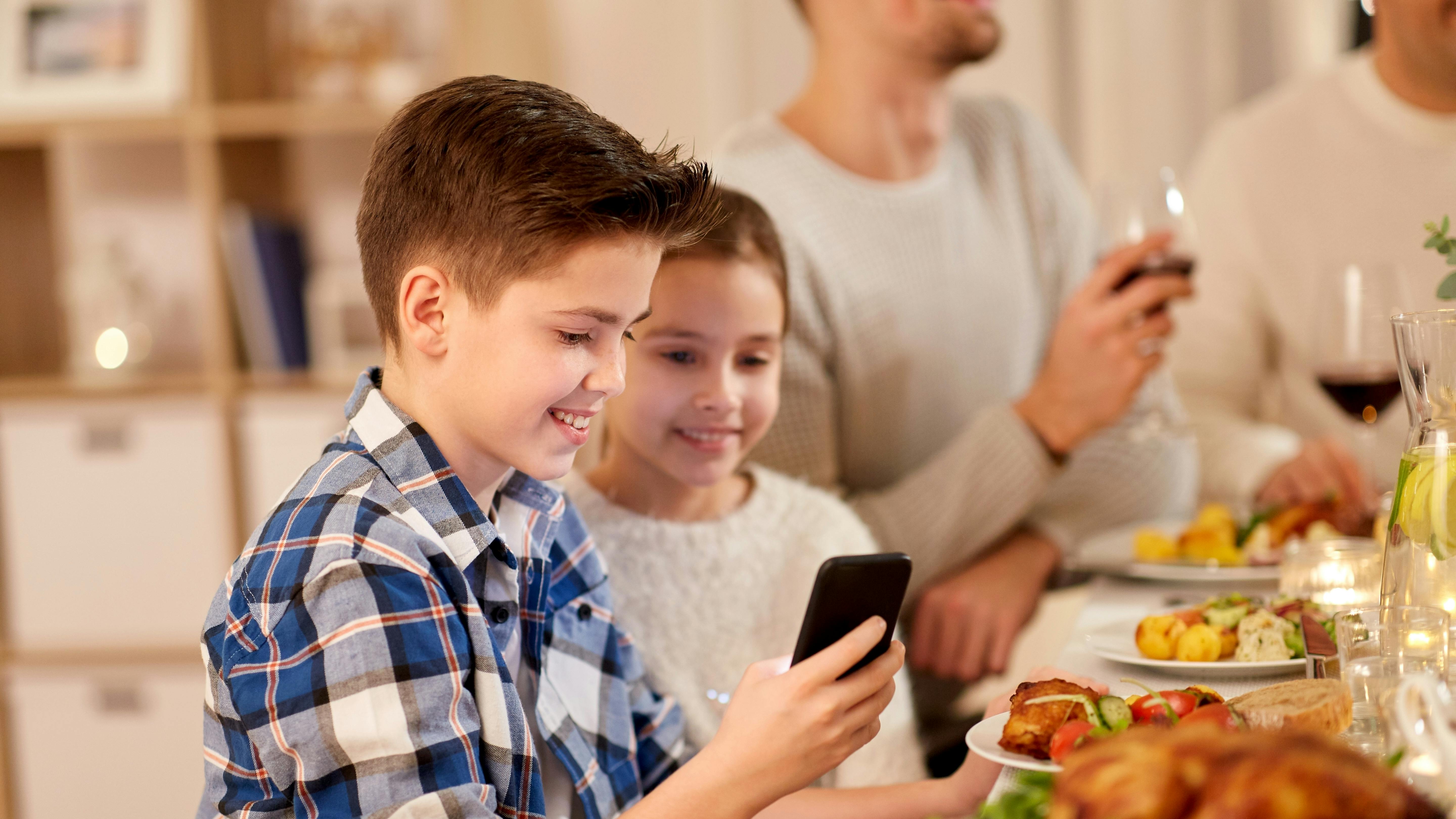 technology, holidays and people concept - happy boy with sister using smartphone having family dinner party at home