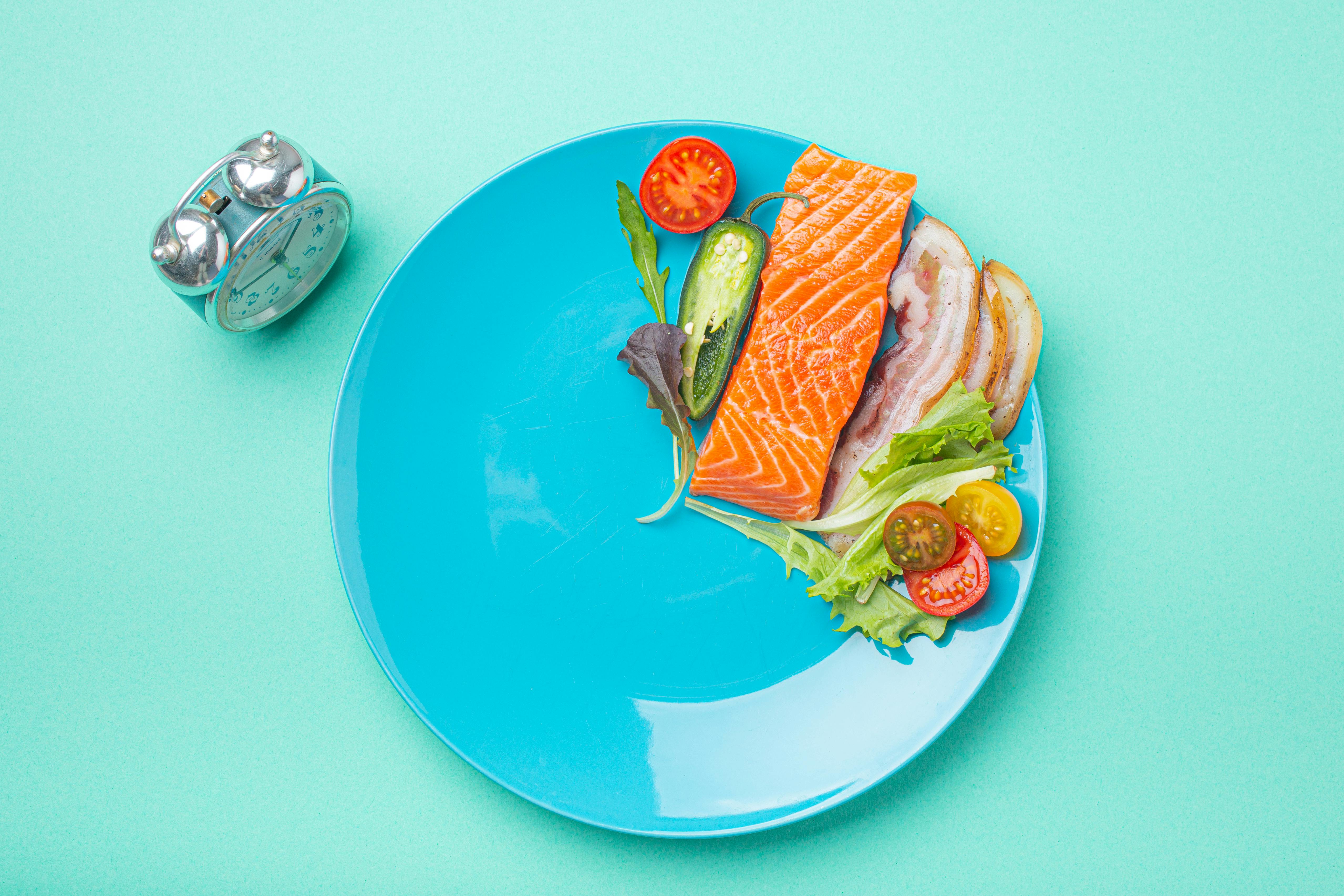 Intermittent fasting low carb hight fats diet concept flat lay, healthy food salmon fish, bacon meat, vegetables and salad on blue plate and clock alarm on blue background top view