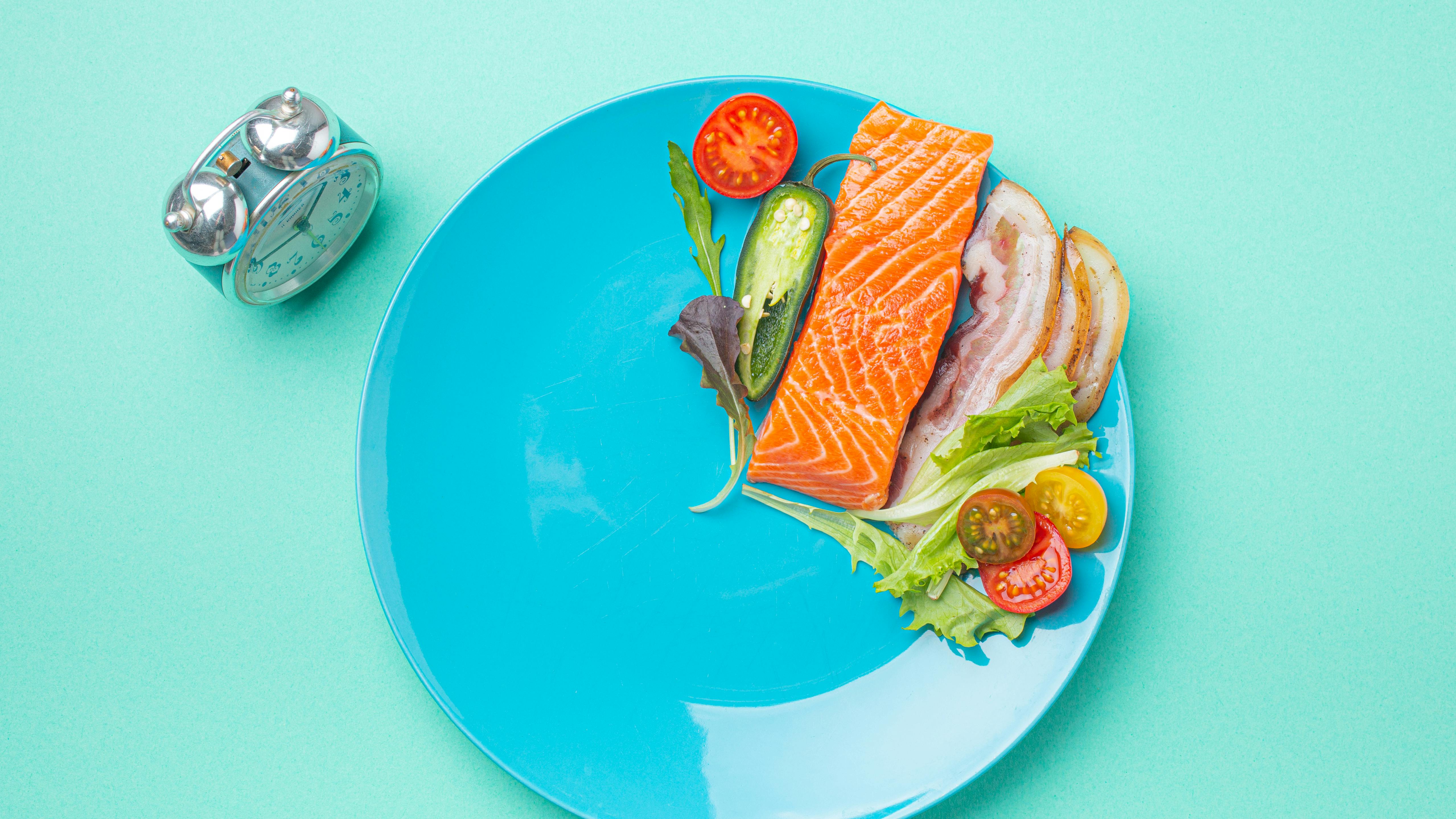 Intermittent fasting low carb hight fats diet concept flat lay, healthy food salmon fish, bacon meat, vegetables and salad on blue plate and clock alarm on blue background top view