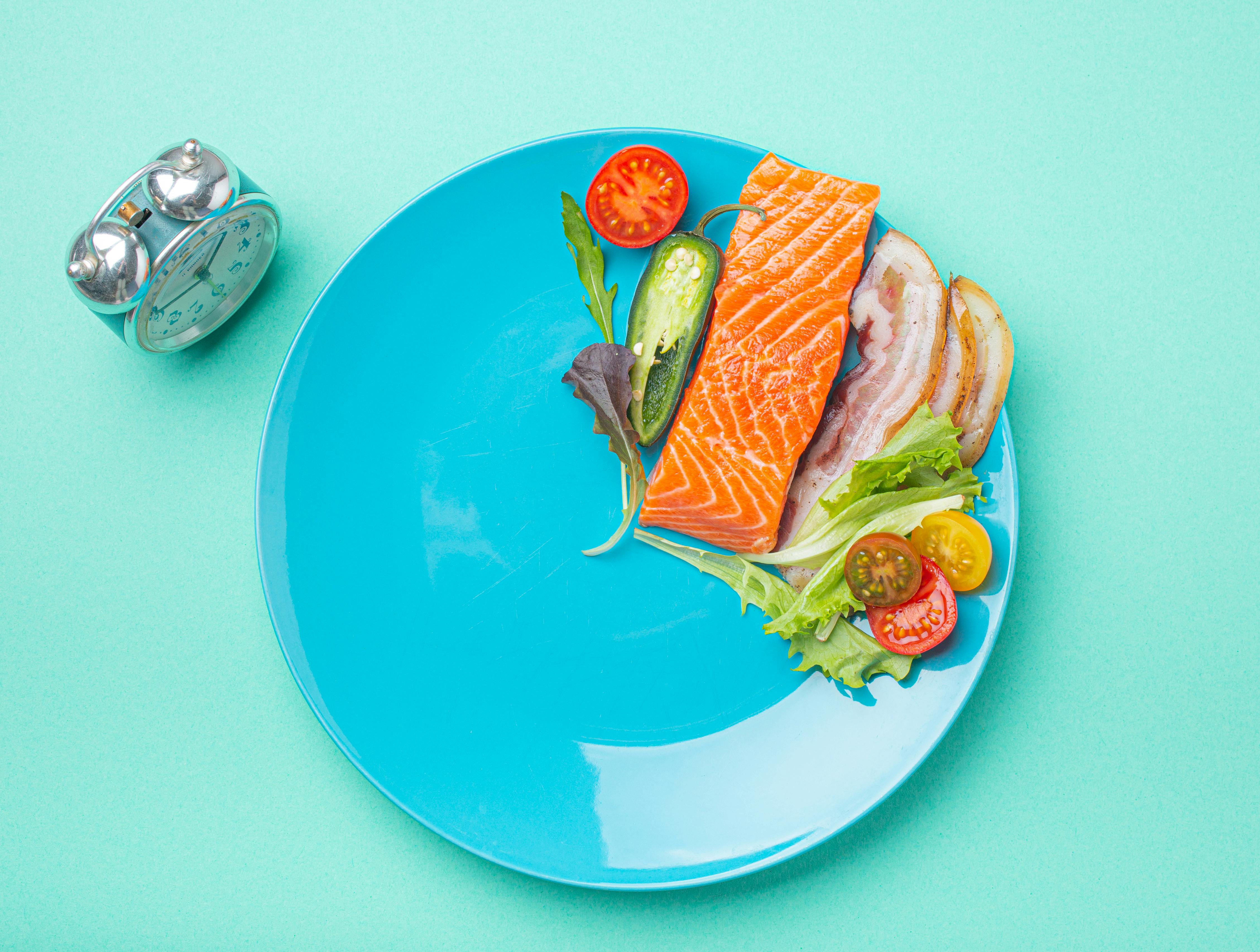 Intermittent fasting low carb hight fats diet concept flat lay, healthy food salmon fish, bacon meat, vegetables and salad on blue plate and clock alarm on blue background top view