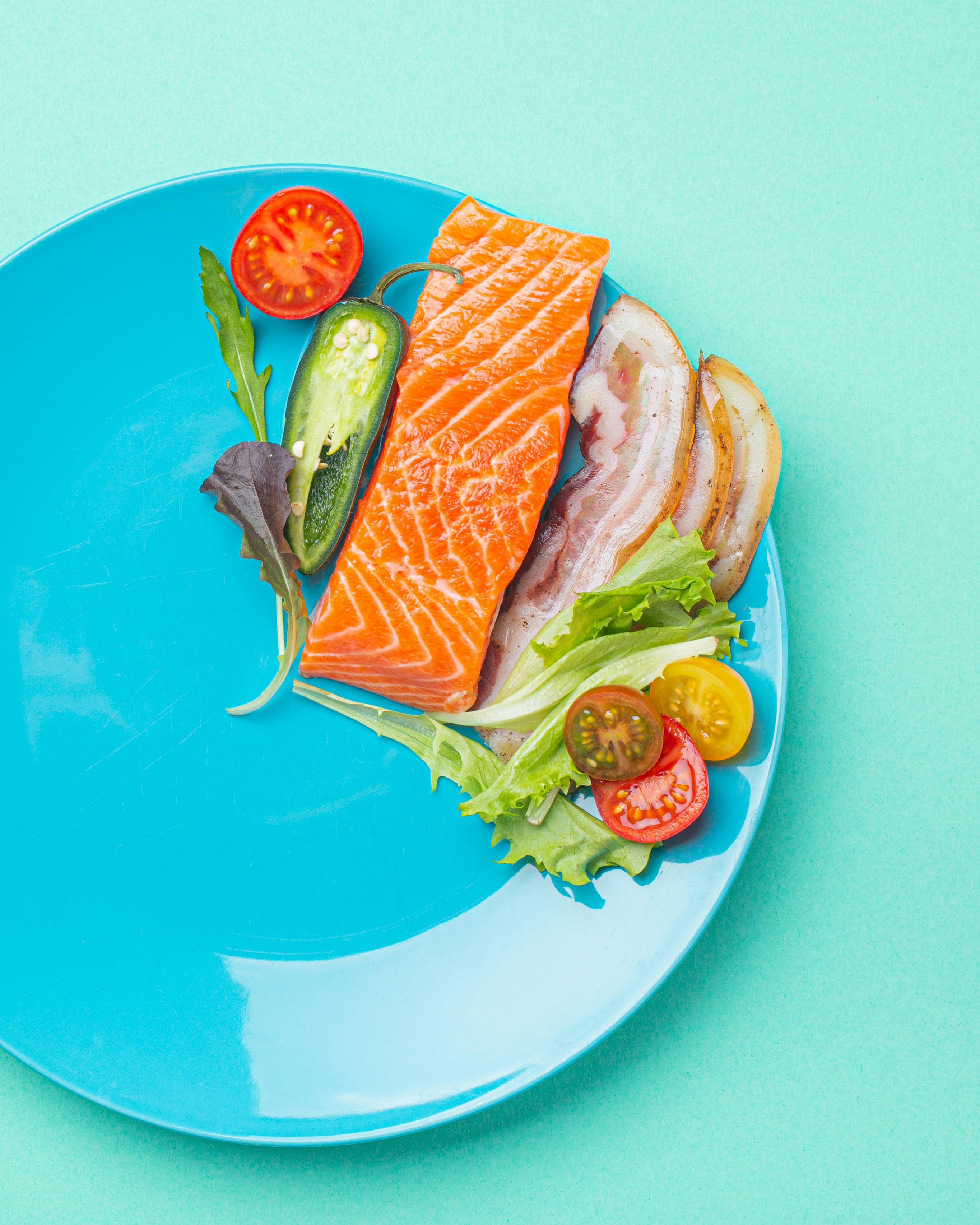 Intermittent fasting low carb hight fats diet concept flat lay, healthy food salmon fish, bacon meat, vegetables and salad on blue plate and clock alarm on blue background top view
