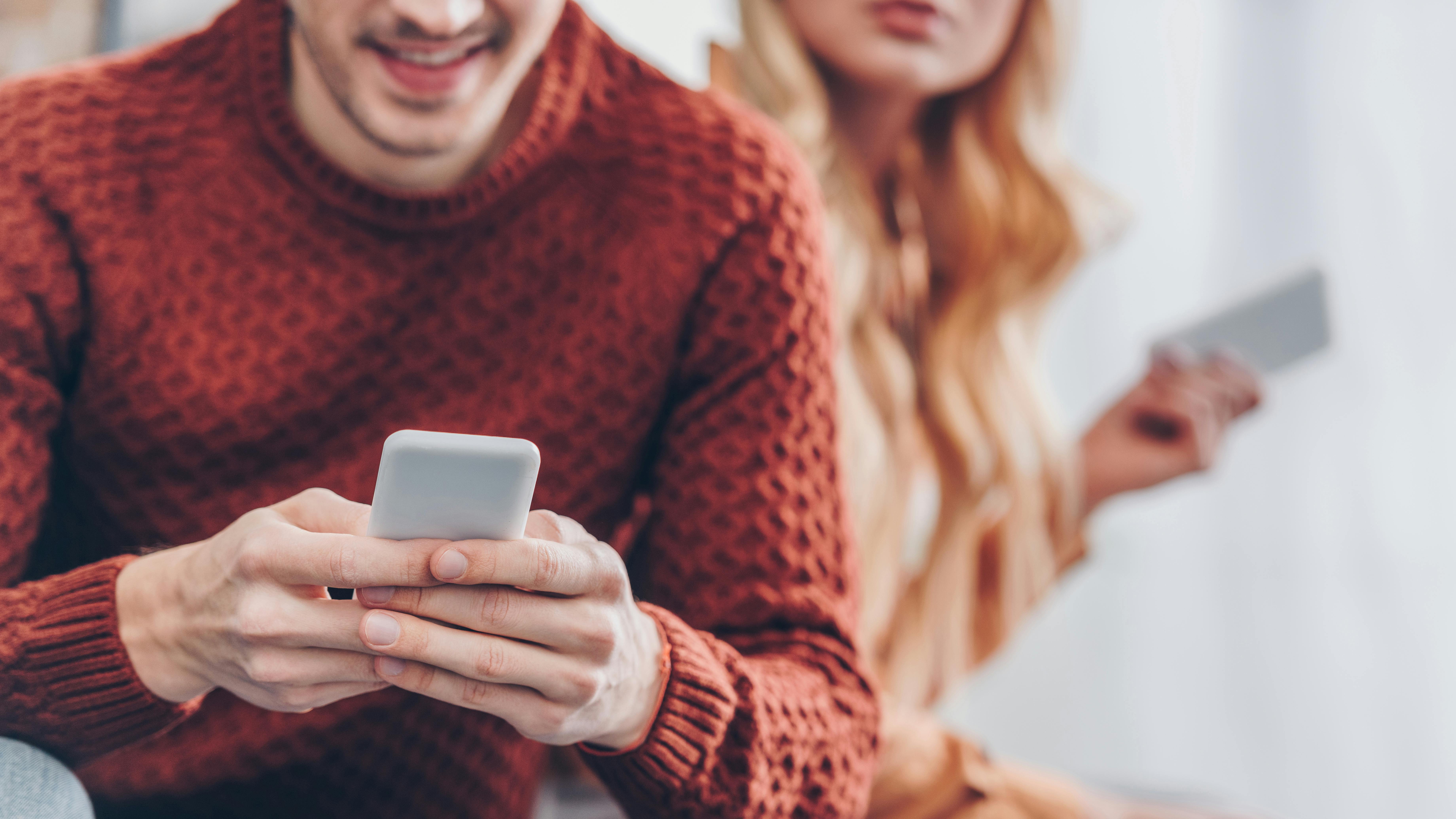 smilende mand sidder med sin mobil, mens en kvinde sidder i baggrunden med sin mobil