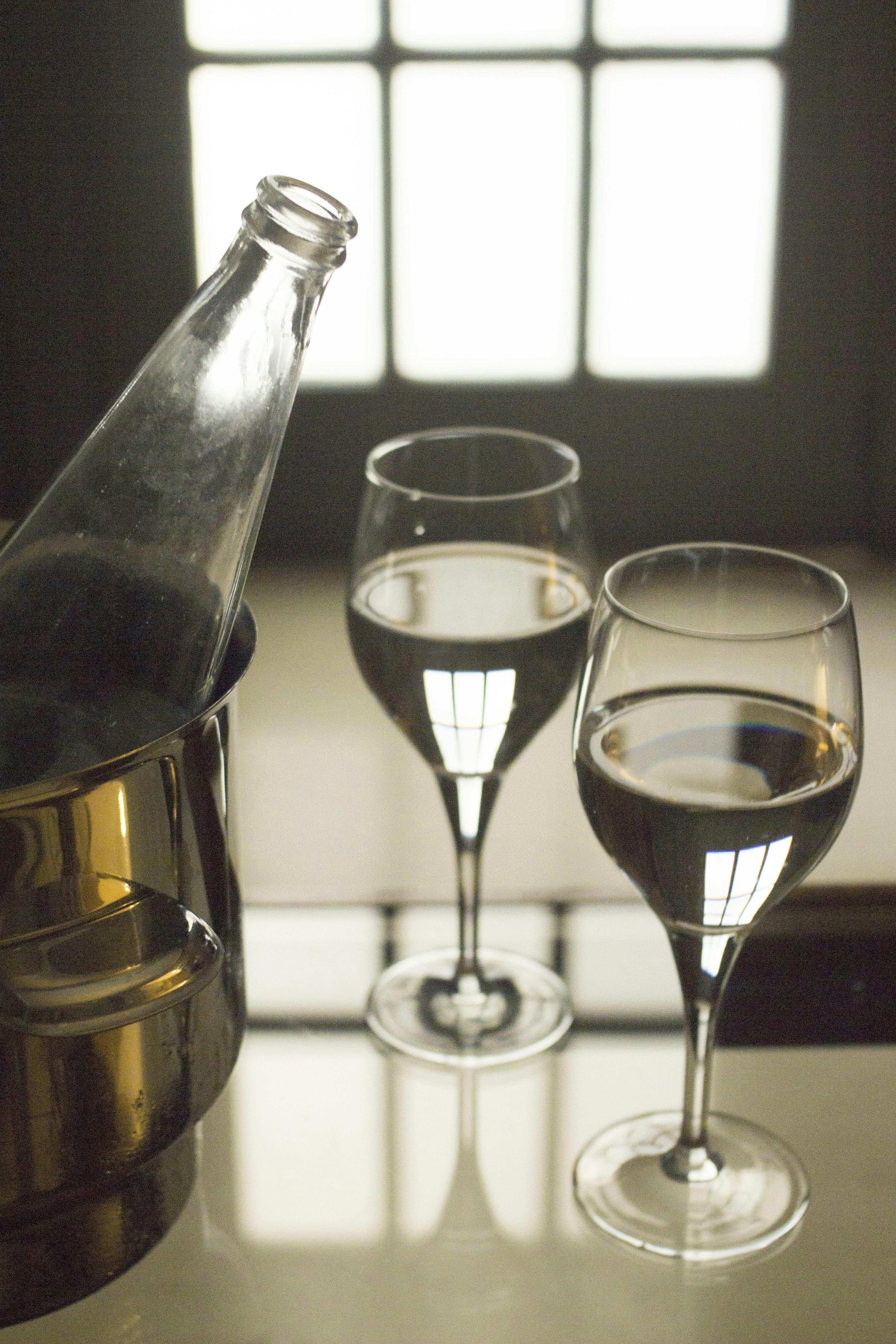 Glass water bottle open, metal ice bucket and two glasses on glass coffee table in room with glass door behind defocused. Monochrome vertical color digital photograph.