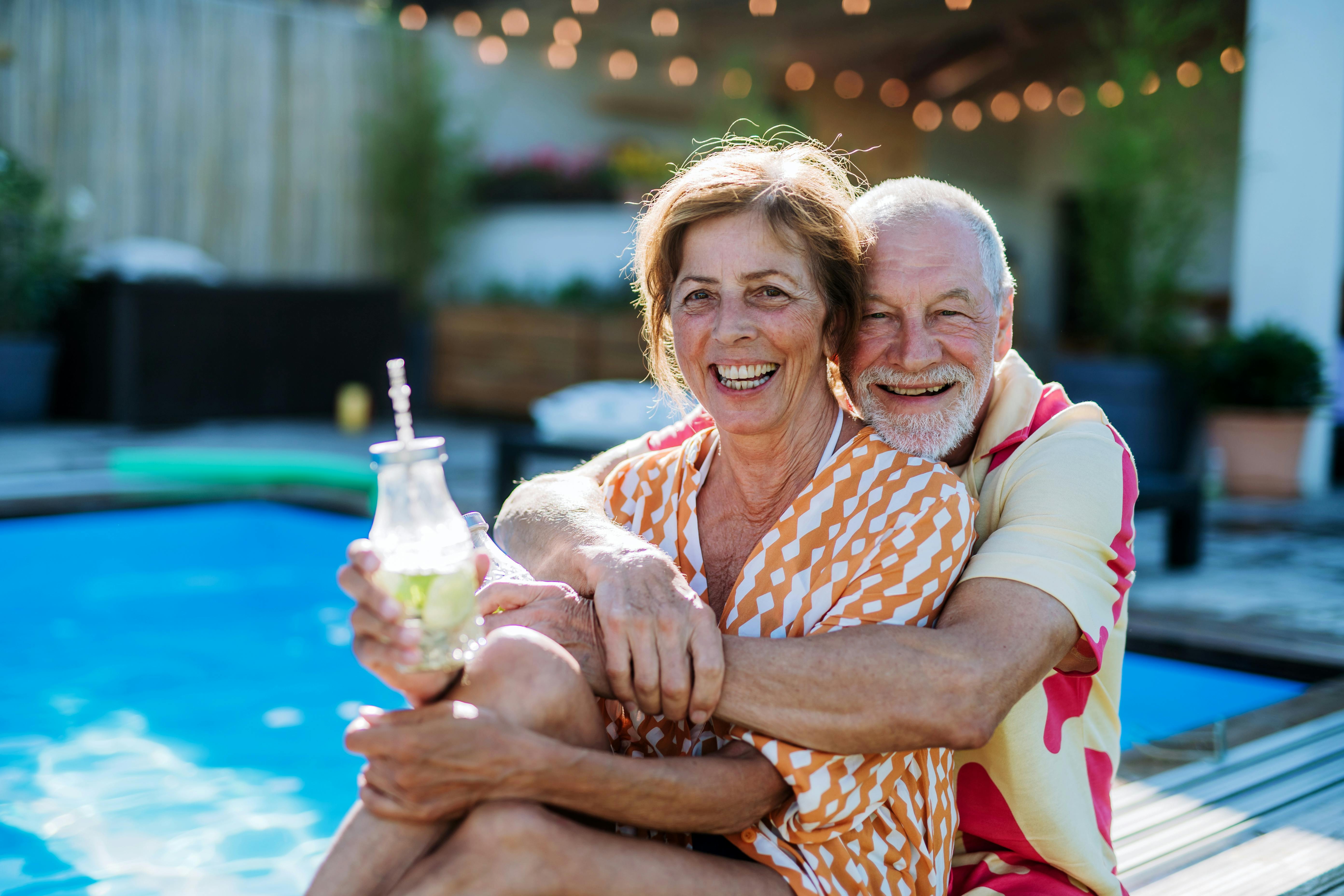 A happy senior couple enjoying drinks when relaxing and sitting by swimming pool in summer.