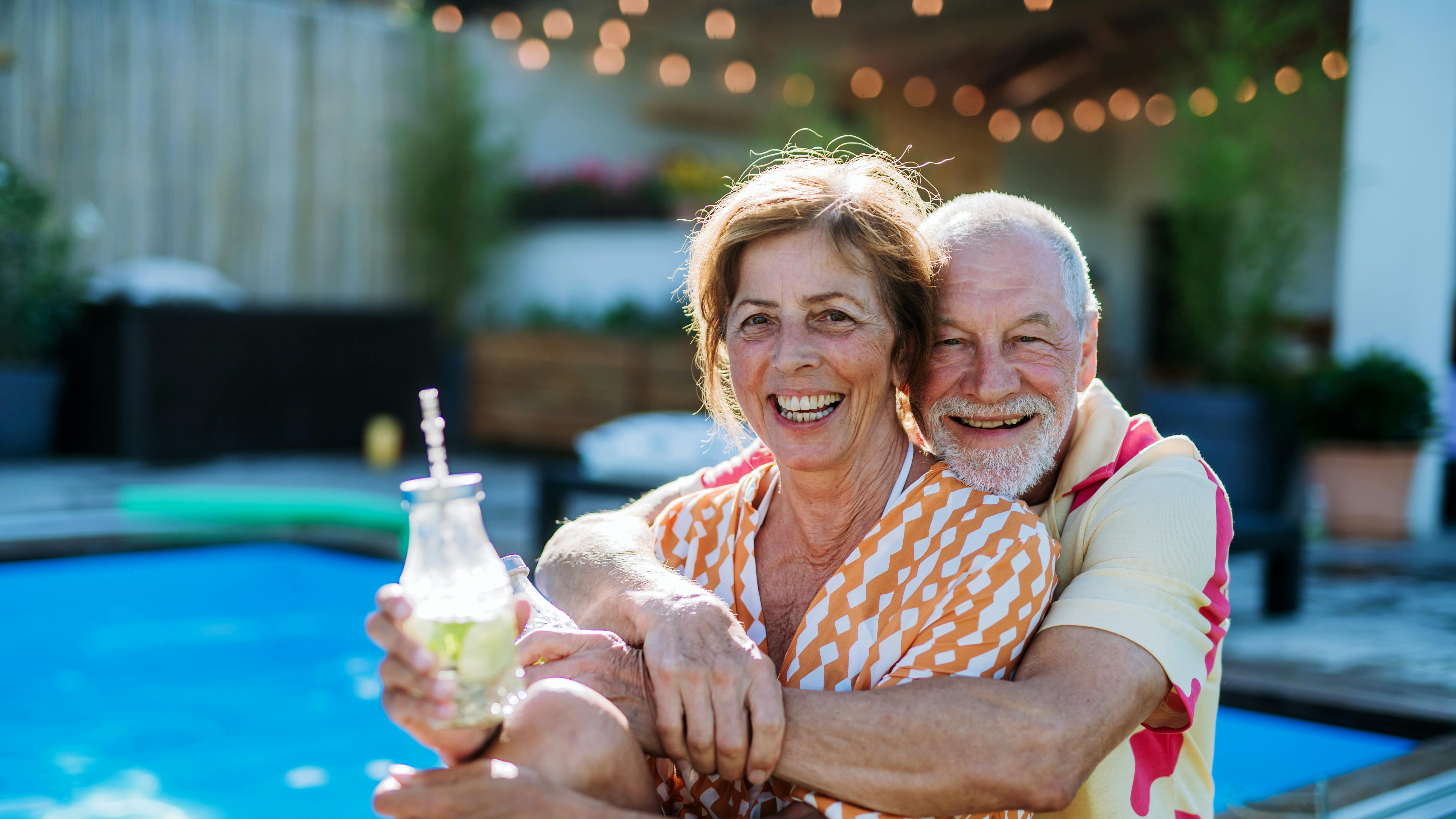 A happy senior couple enjoying drinks when relaxing and sitting by swimming pool in summer.
