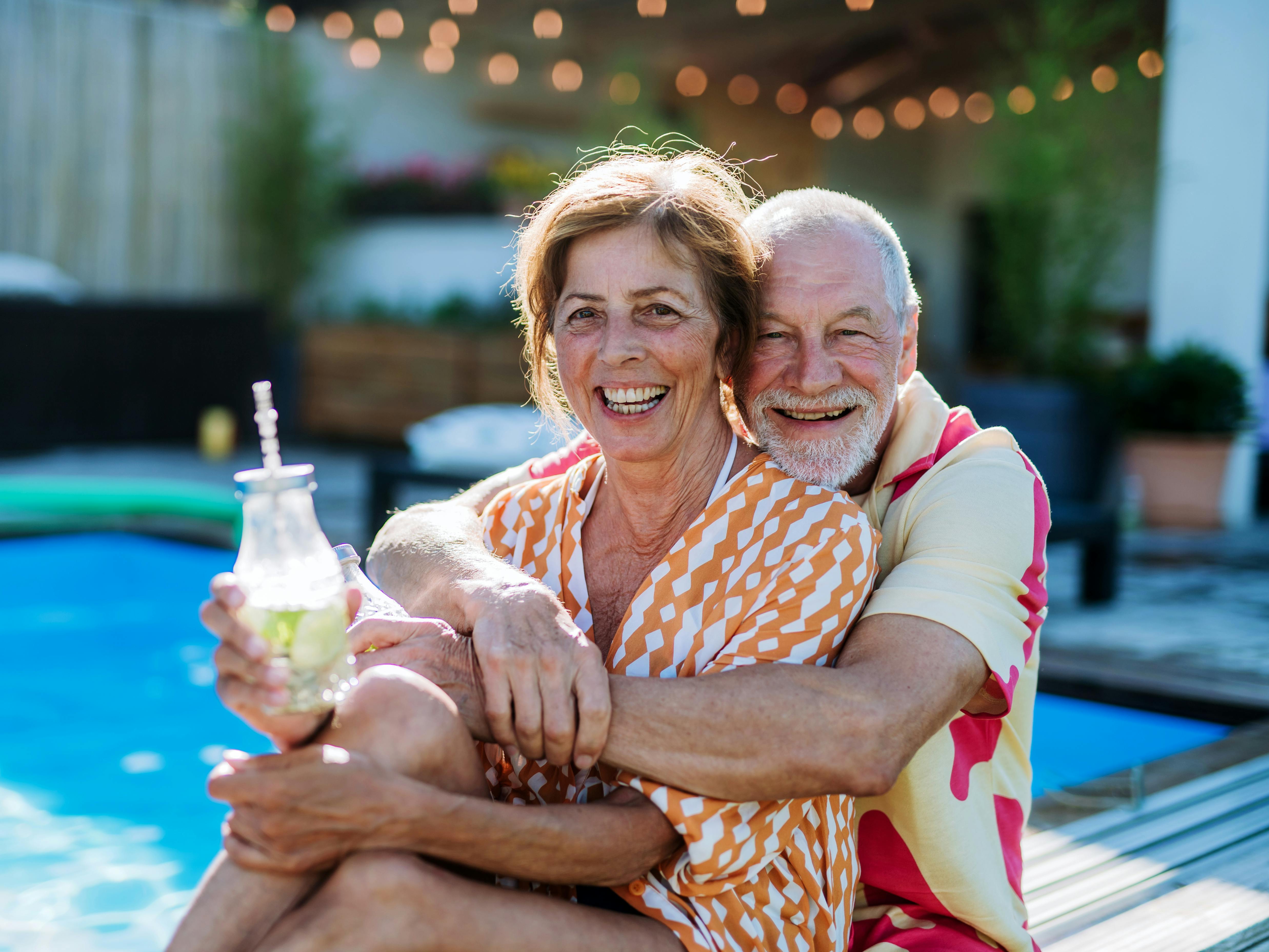 A happy senior couple enjoying drinks when relaxing and sitting by swimming pool in summer.