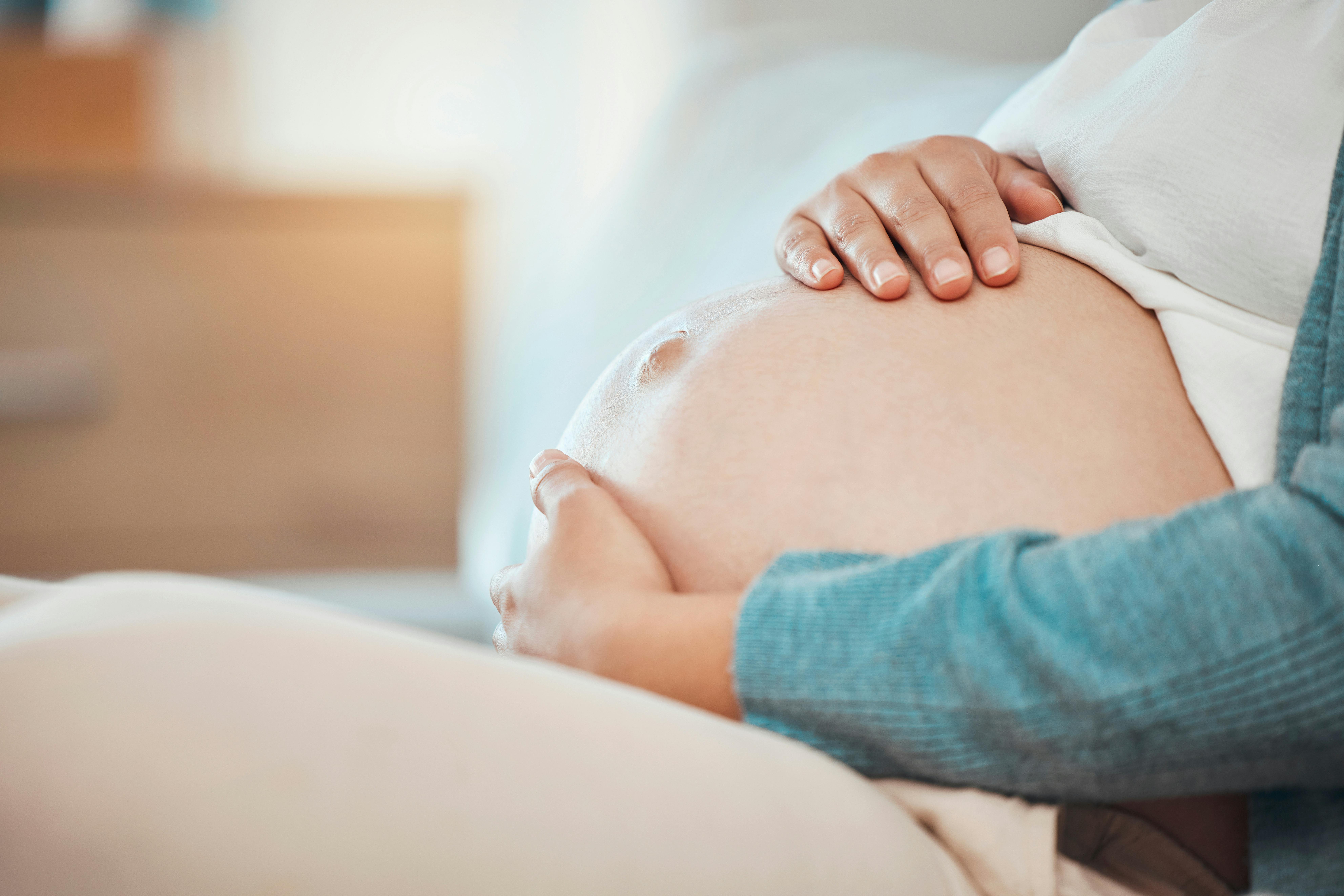 Hands, stomach and pregnant with a woman sitting in the bedroom of her home alone while touching her belly. Mother, love and baby with a female parent holding her tummy while on the bed to relax.