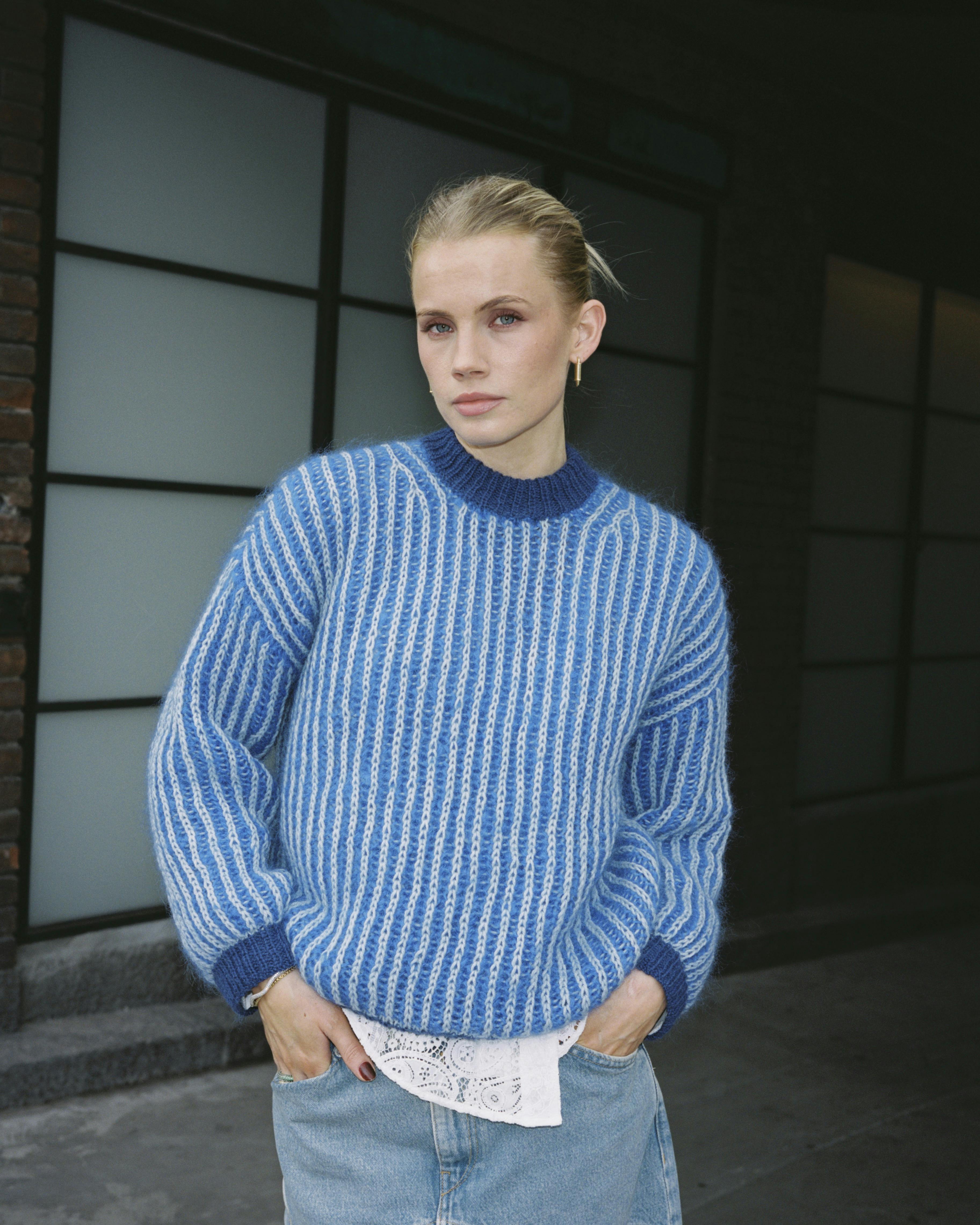 Strikkeopskrift på blå patentsweater, hvor kvinden står med hænderne i nederdelen.