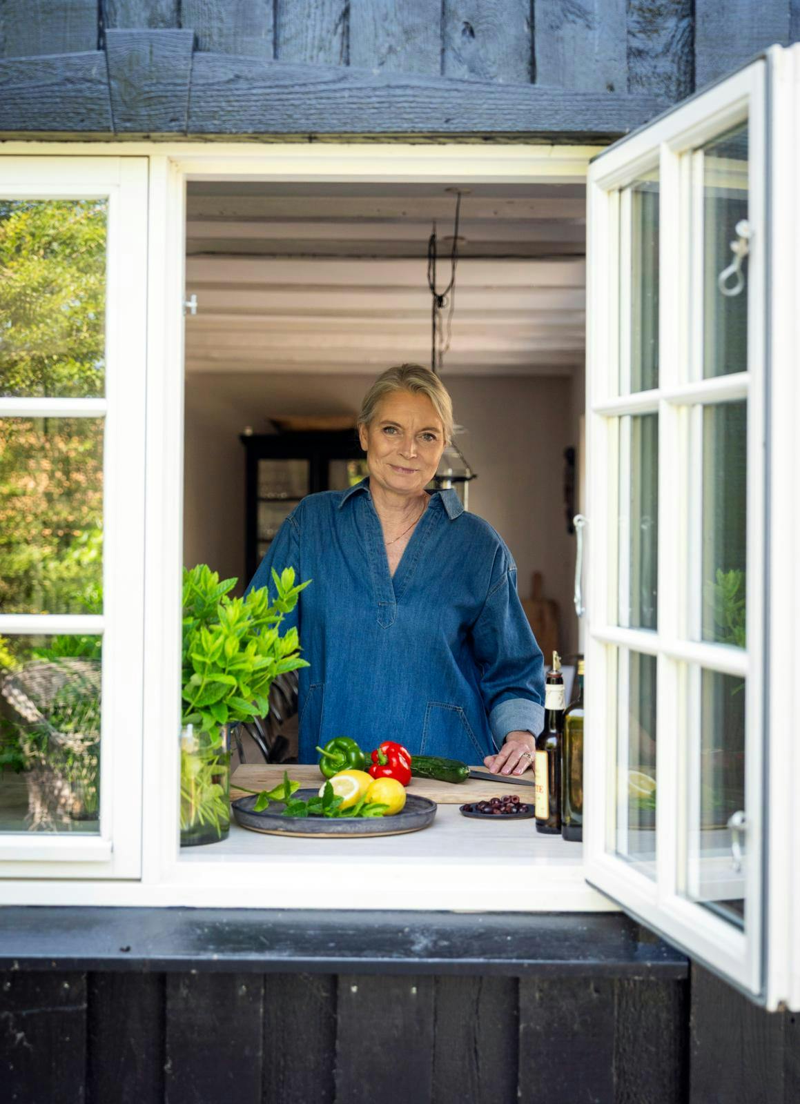 Kvinde i et køkken, der kigger ud af et vindue. Foran hende ligger et spækbræt med skårne grøntsager.