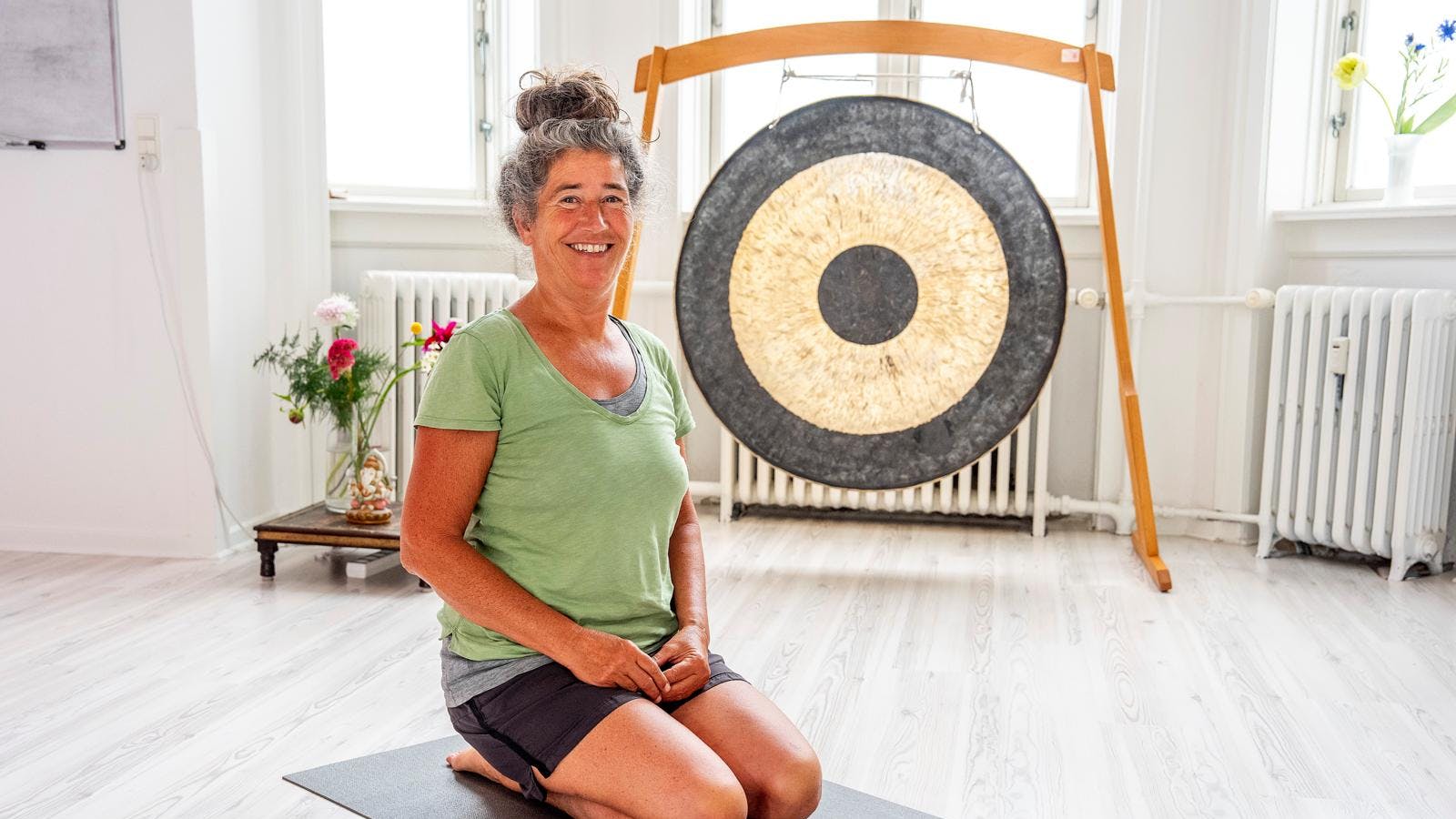 En kvinde, der sidder på knæ på gulvet i yogatøj og smiler. Bagved ses en sort og guldfarvet  gong-gong.