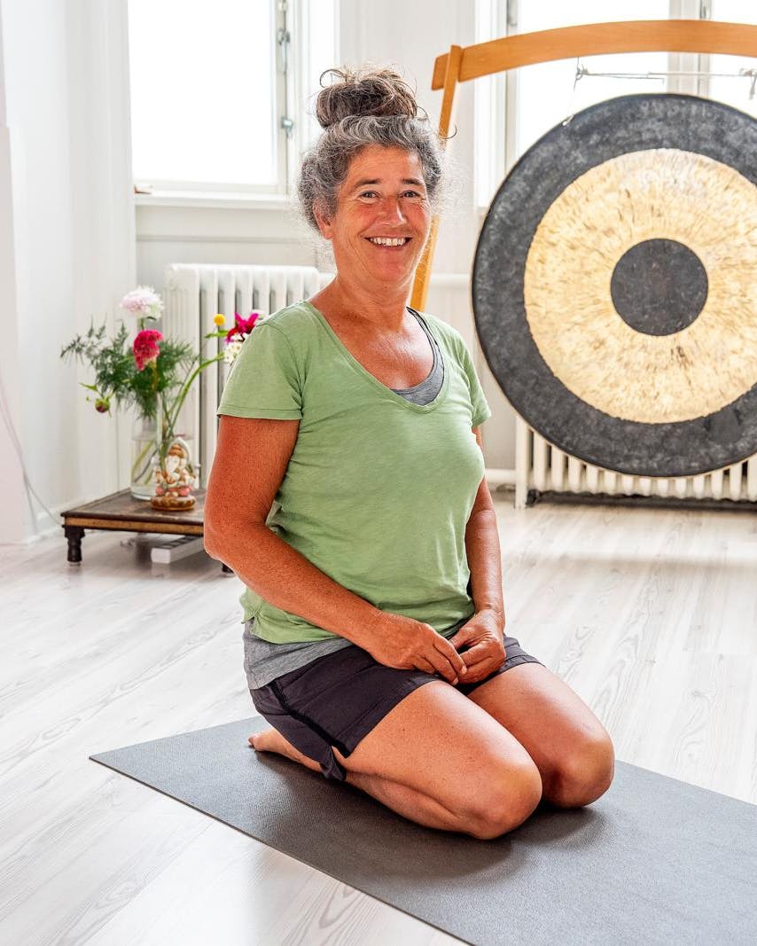 En kvinde, der sidder på knæ på gulvet i yogatøj og smiler. Bagved ses en sort og guldfarvet gong-gong.