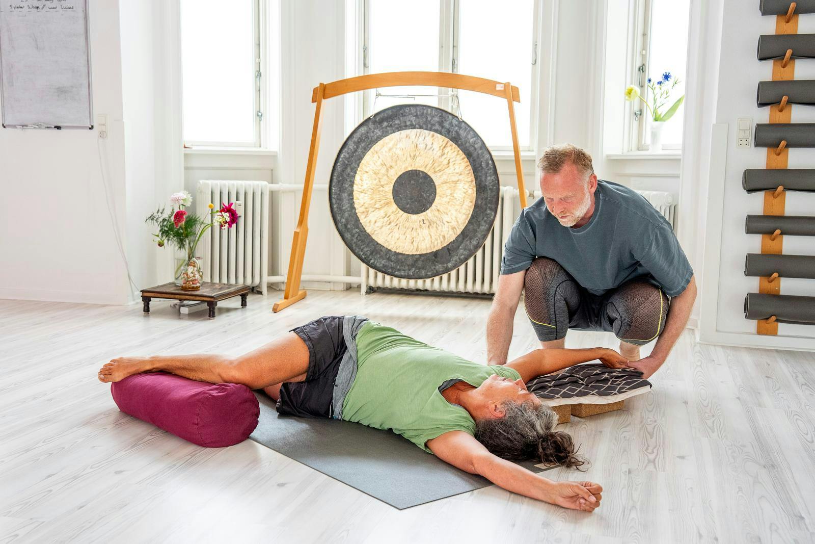 Kvinde, der ligger på gulvet og dyrker yoga, mens en instruktør retter på hendes ben. 