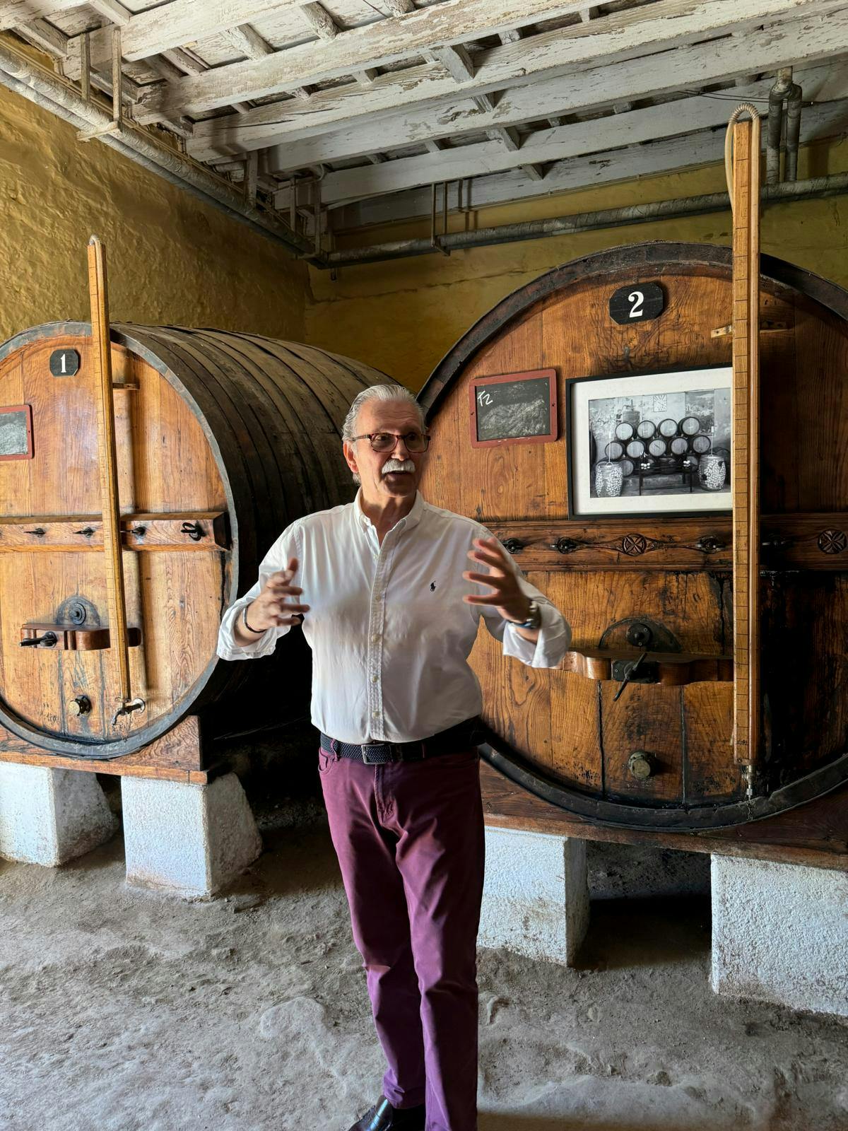 En mand med løftede arme, der står foran to store vintønder og snakker.