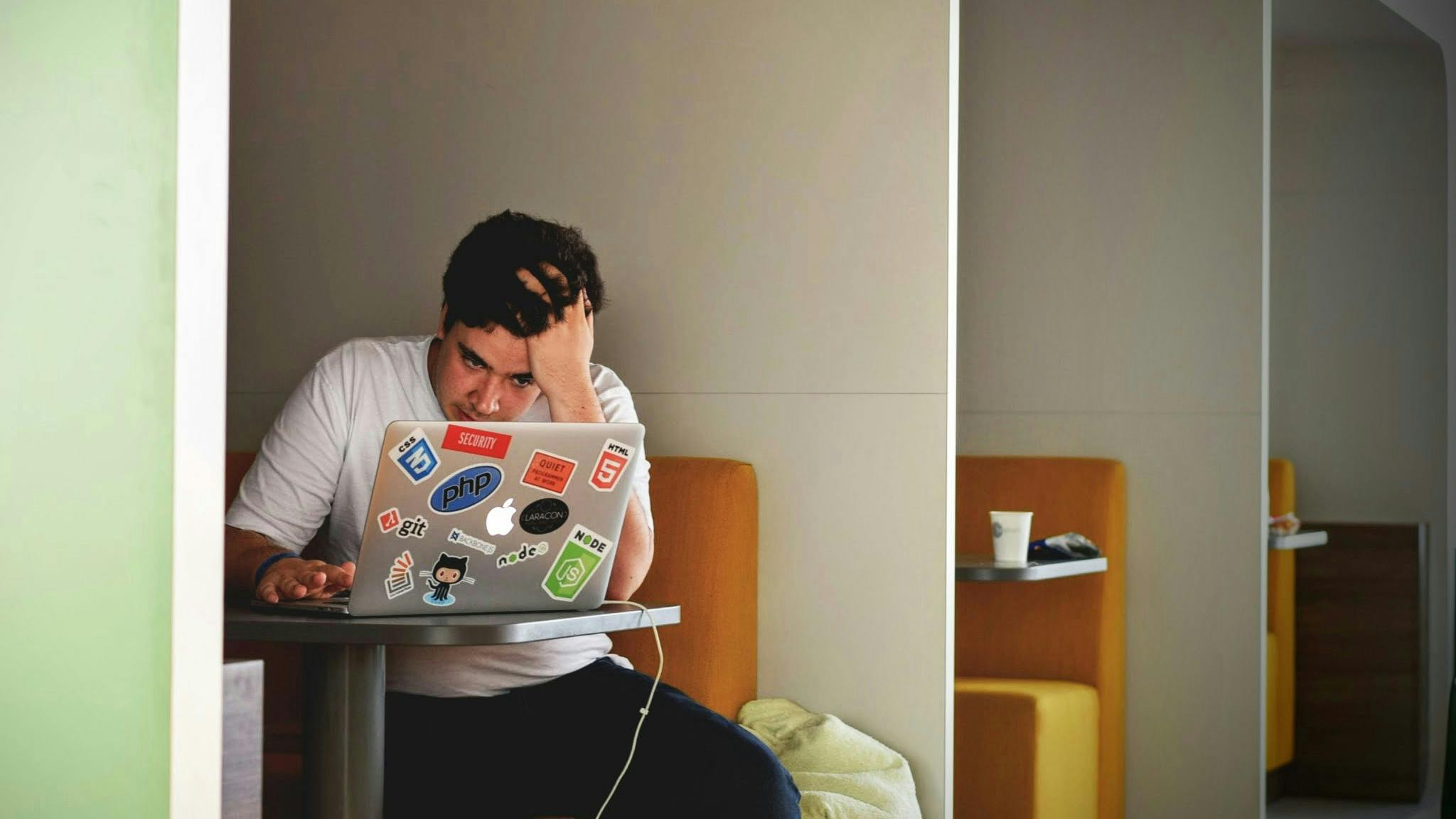 En ung mand, der sidder på en cafe med en pc foran sig og tager sig til håret.