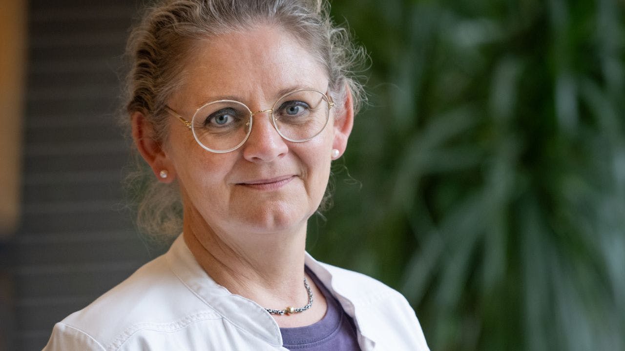 Læge med briller og kittel smiler foran sløret plante