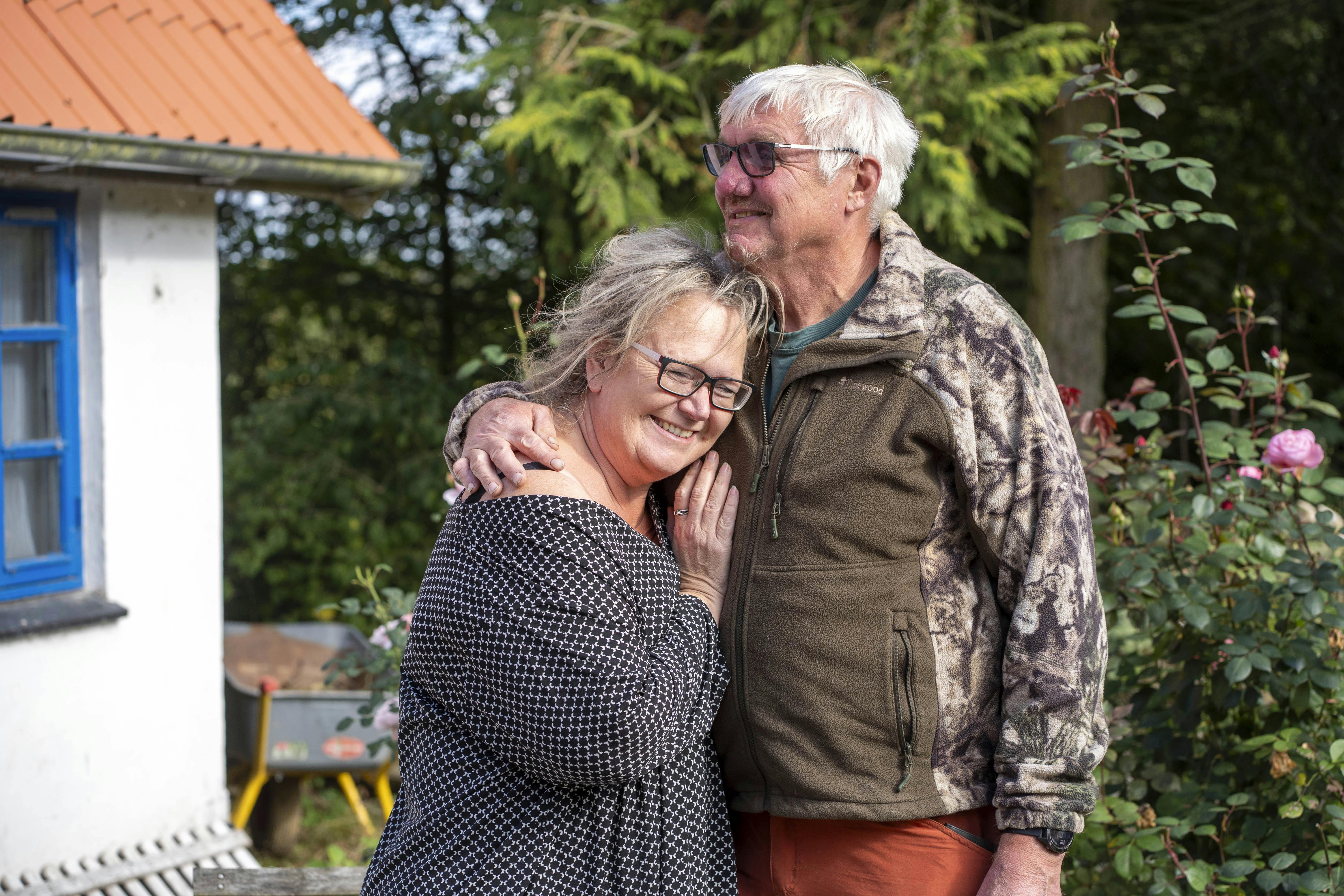Ældre par krammer foran et hvidt hus med blå vinduer