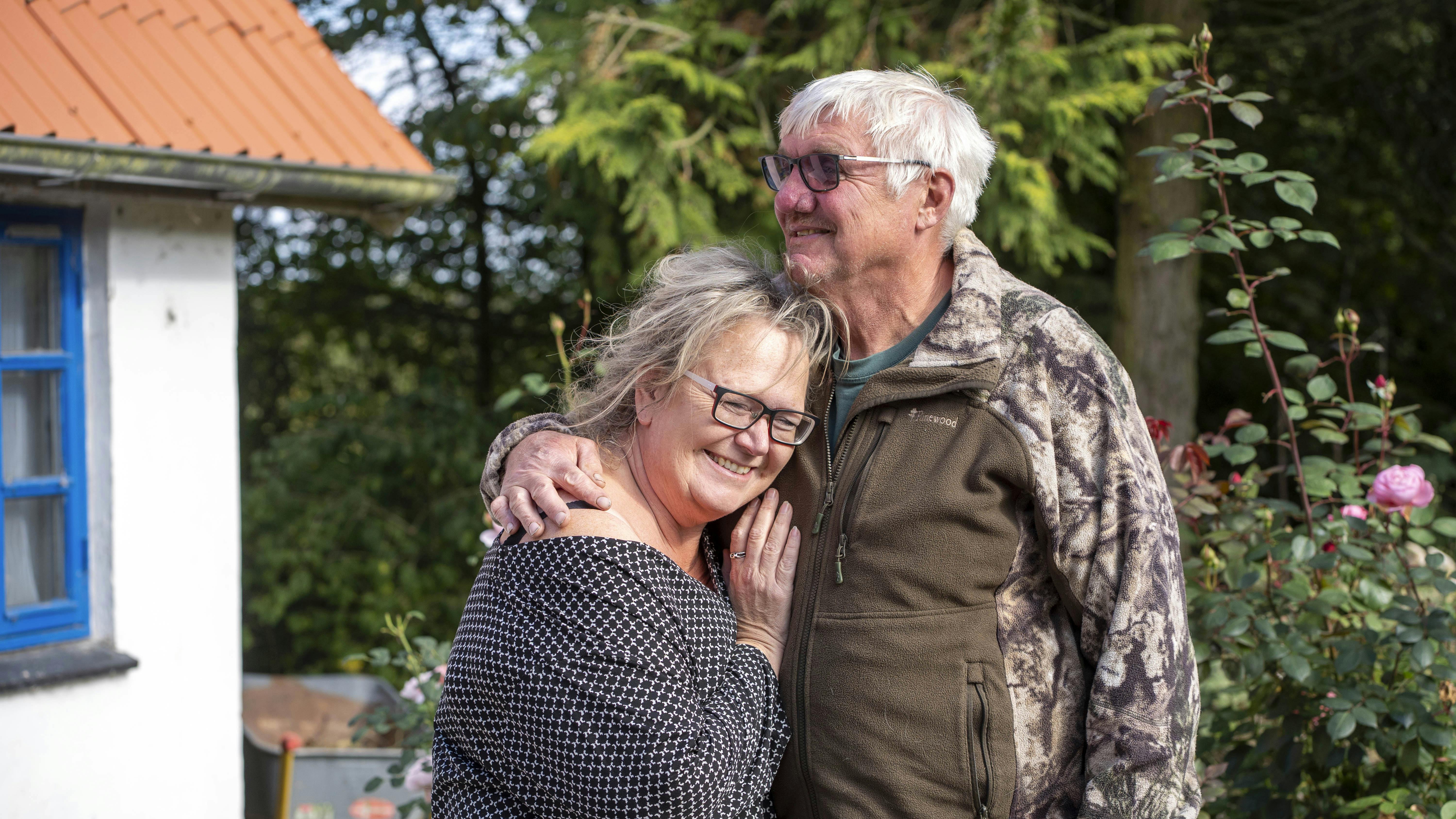 Ældre par krammer foran et hvidt hus med blå vinduer