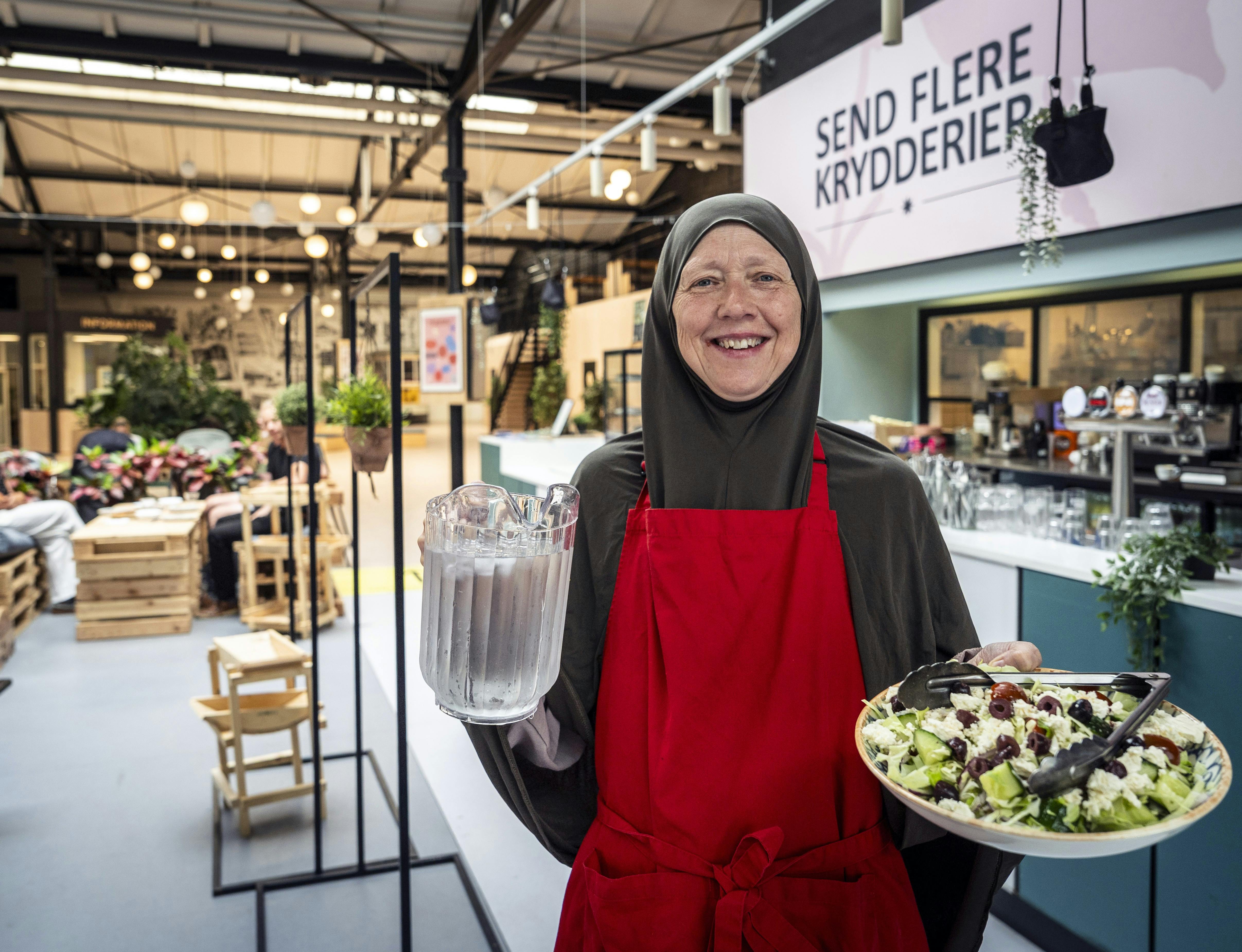 Liselotte står med et fad og en kande og er en af de frivillige på Send flere krydderier