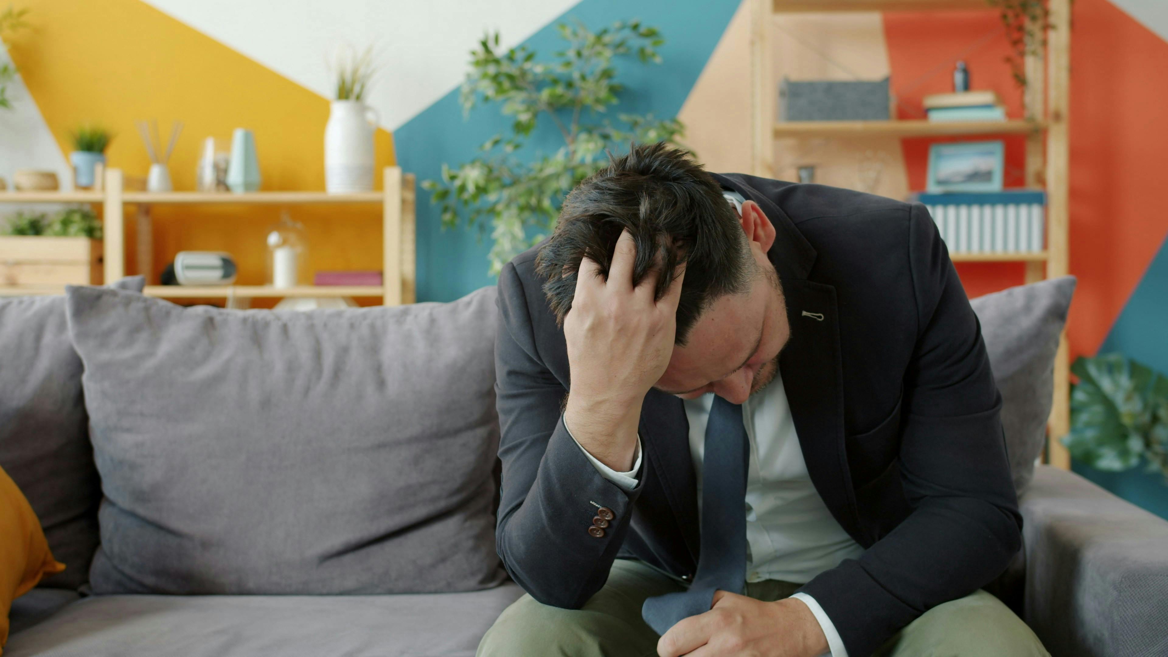 En mand i jakkesæt, der sidder i en sofa med bøjet hoved og ser nedtrykt ud.