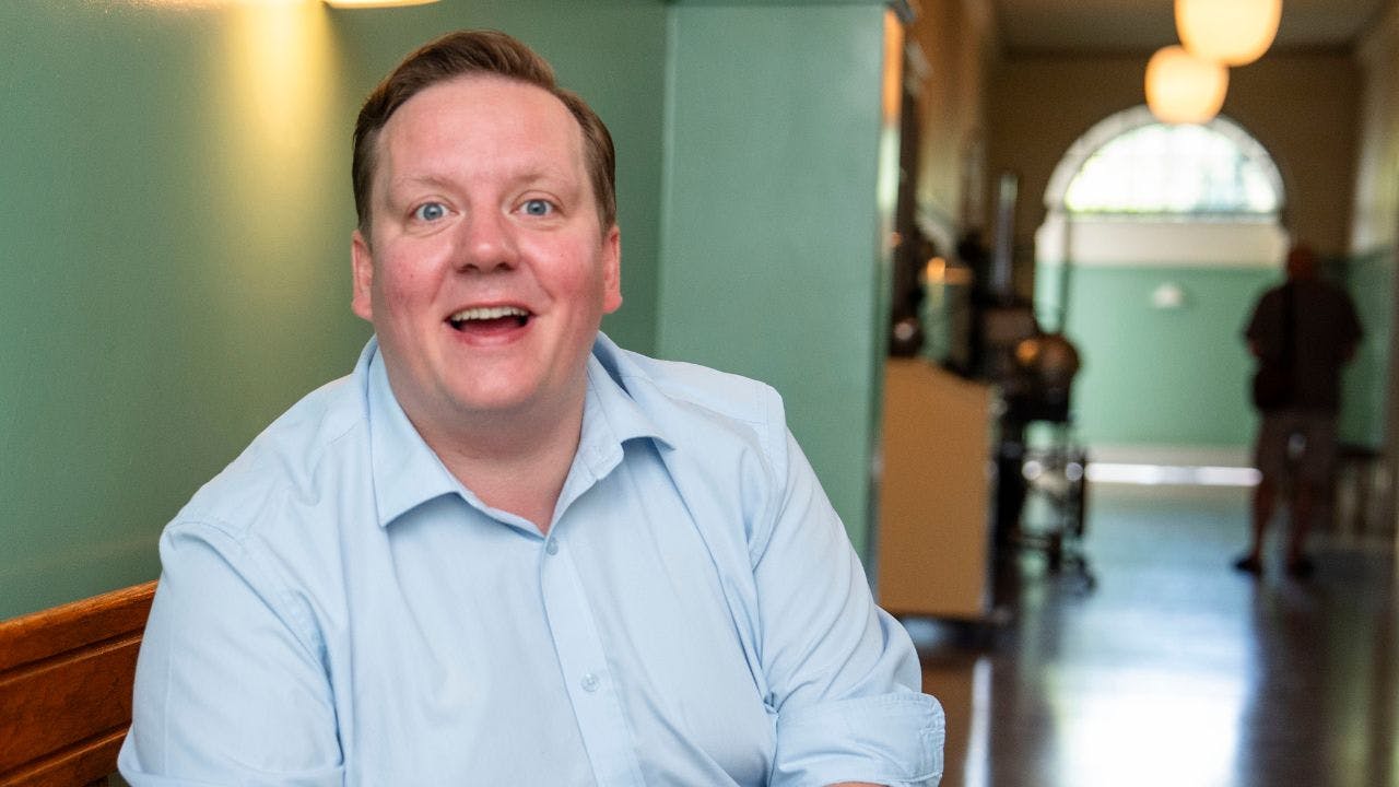 Mand i blå skjorte sidder ned og smiler stort på en hospitalsgang på et filmset