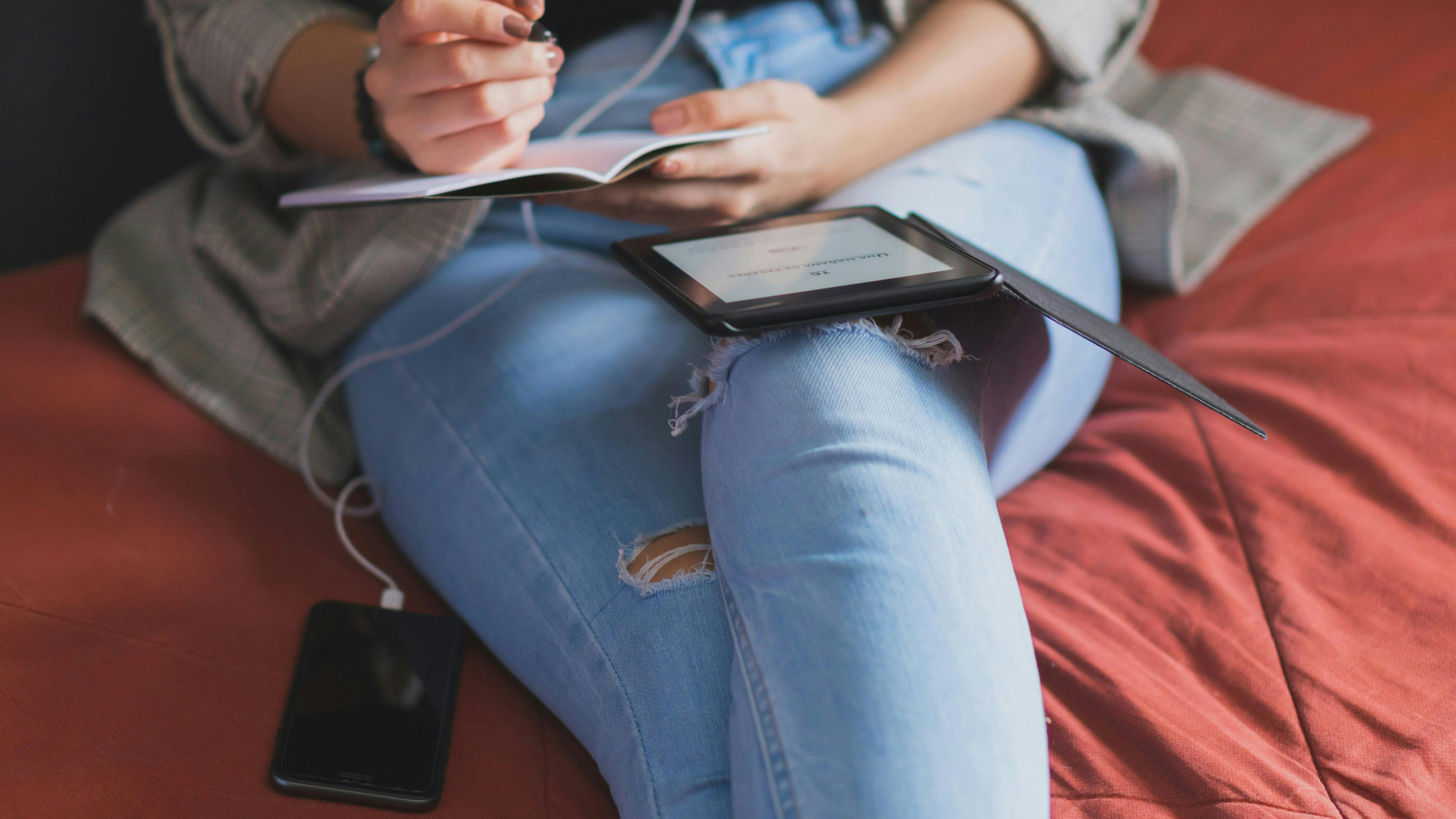 Krydsede ben i slidte jeans og en overkrop, der sidder i en seng. Personen sidder med ipad og iphone og skriver.