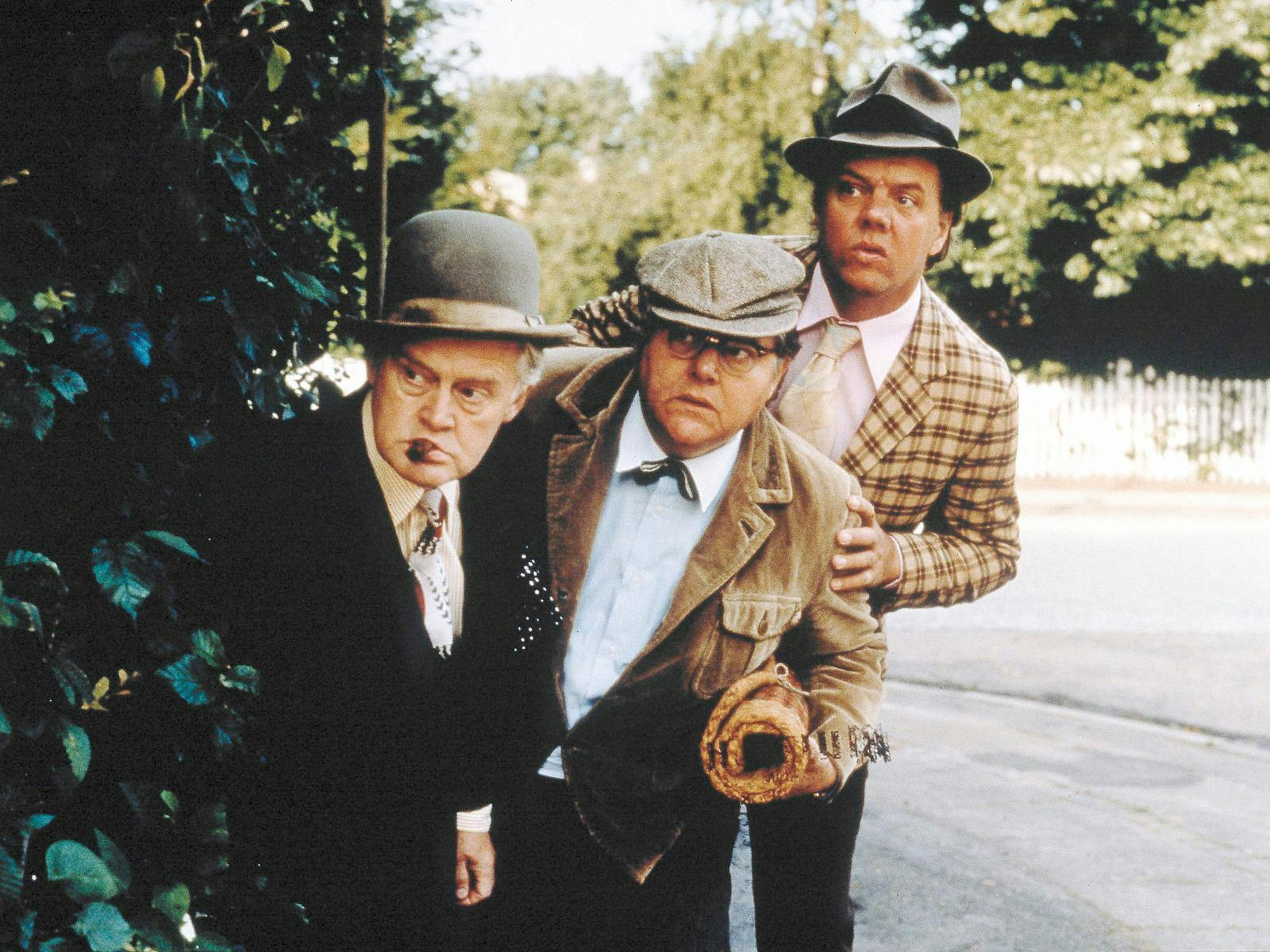 Tre midaldrende mænd - Olsen-banden - der gemmer sig bag et træ og kigger efter noget. 