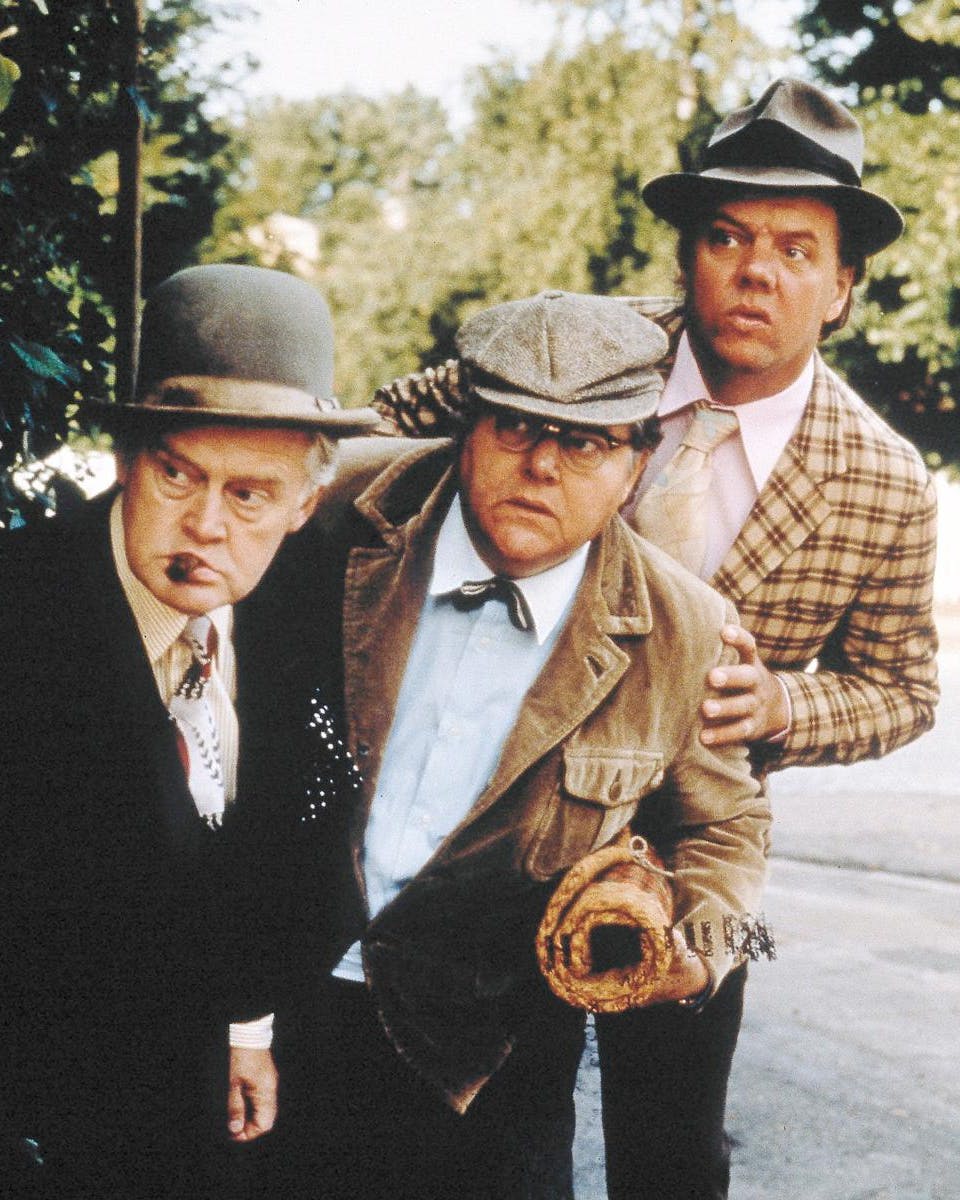 Tre midaldrende mænd - Olsen-banden - der gemmer sig bag et træ og kigger efter noget.