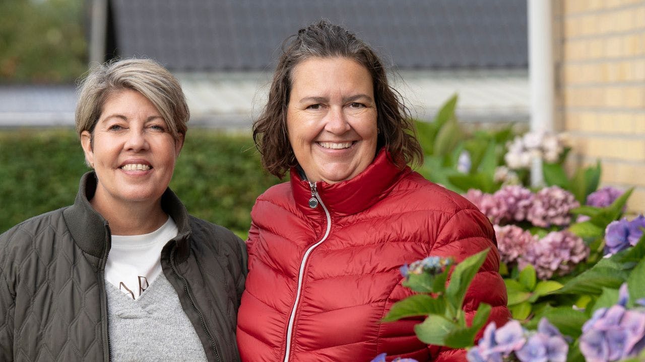 To kvinder står og smiler ved siden af en blomsterbusk