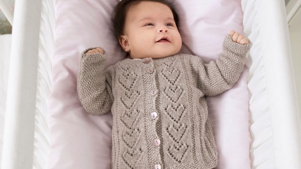 Baby ligger i seng med strikket cardigan på