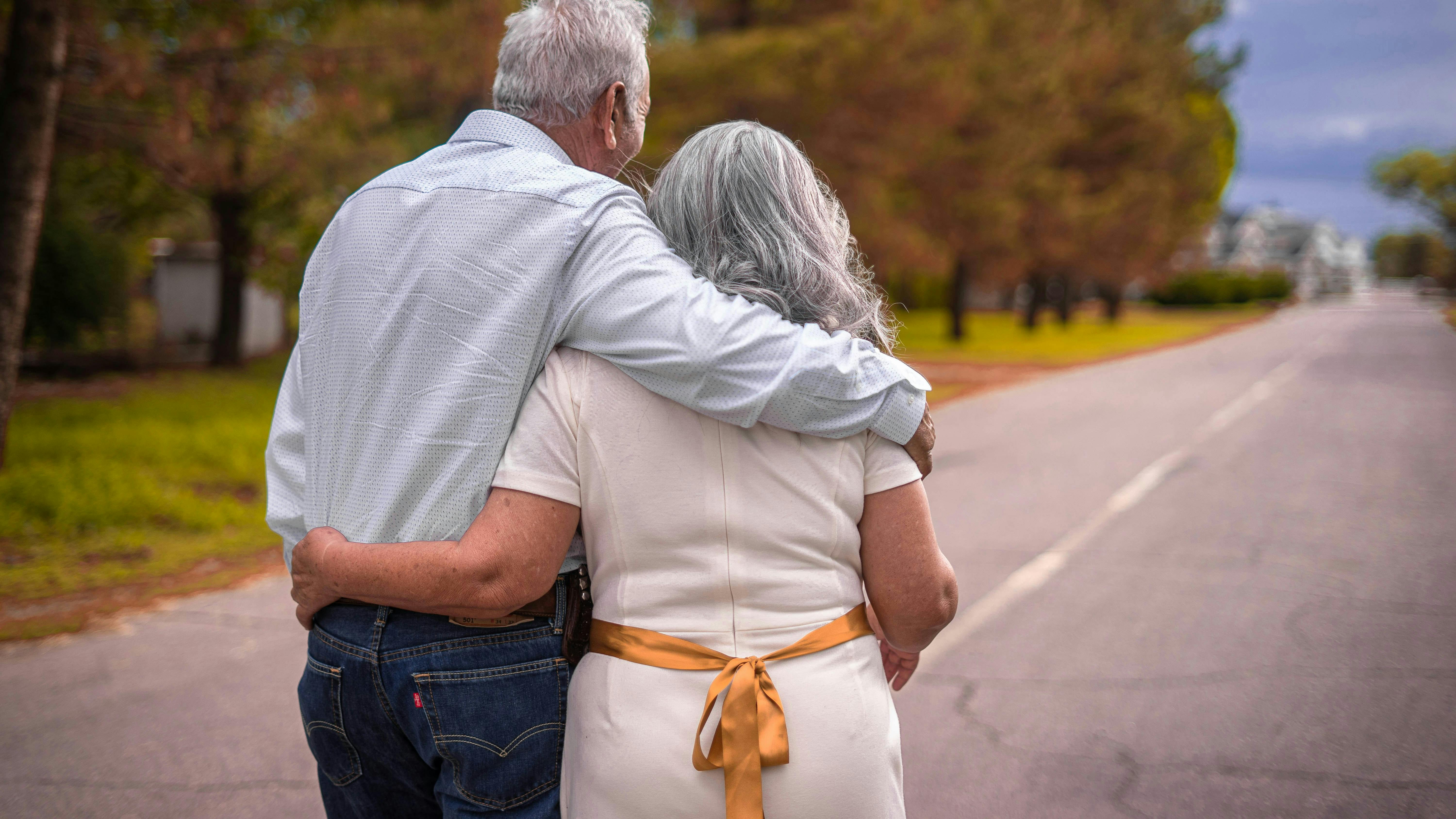 Ældre par med gråt hår, der står midt på en vej med armene om hinanden.