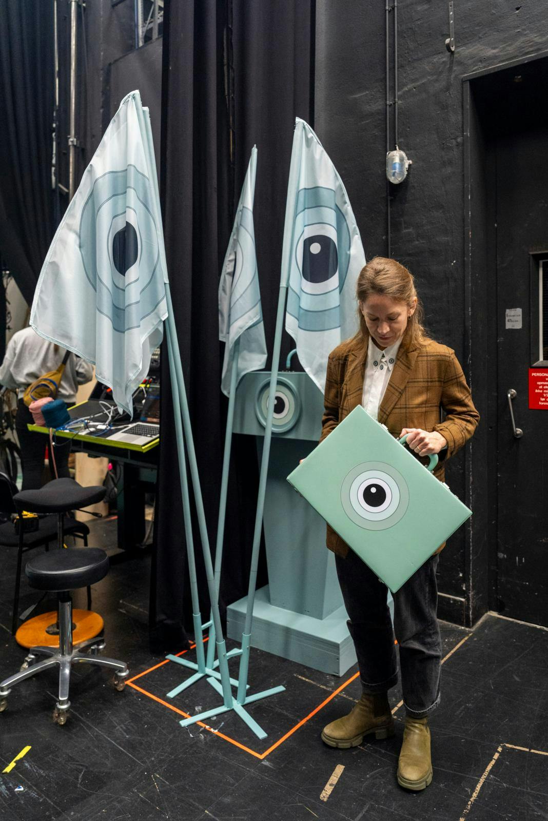 En kvinde, der står med en turkis kuffert med et øje på og nogle flag i baggrunden med samme logo.