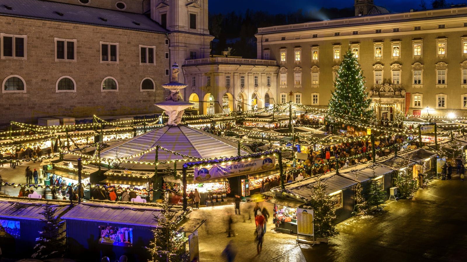 En stor plads omgivet af gamle bygninger set ved aftentid. Det hele er lyst op, og de er juletræ i midten.