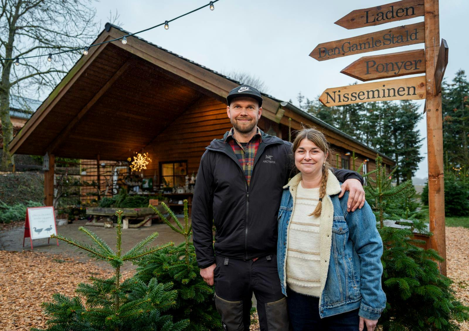 Et par, der står med armene om hinanden foran nogle juletræer og en træhytte.
