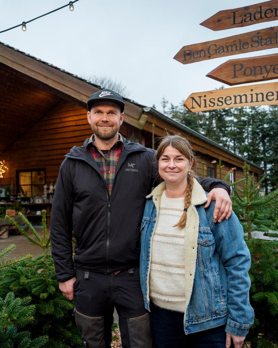 Et par, der står med armene om hinanden foran nogle juletræer og en træhytte.
