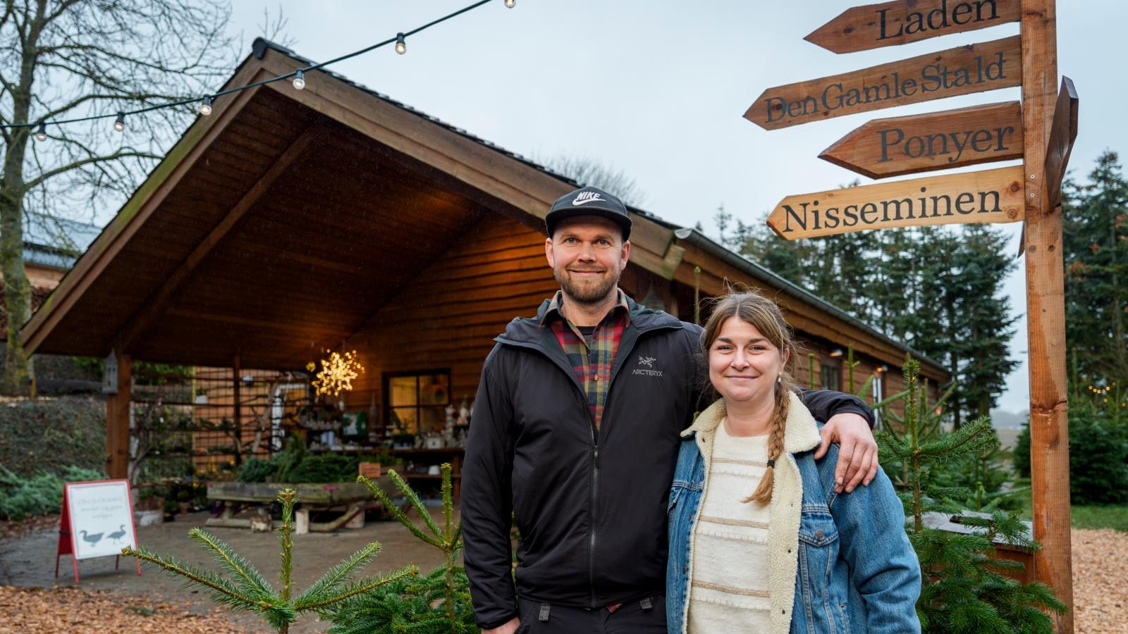 Et par, der står med armene om hinanden foran nogle juletræer og en træhytte.