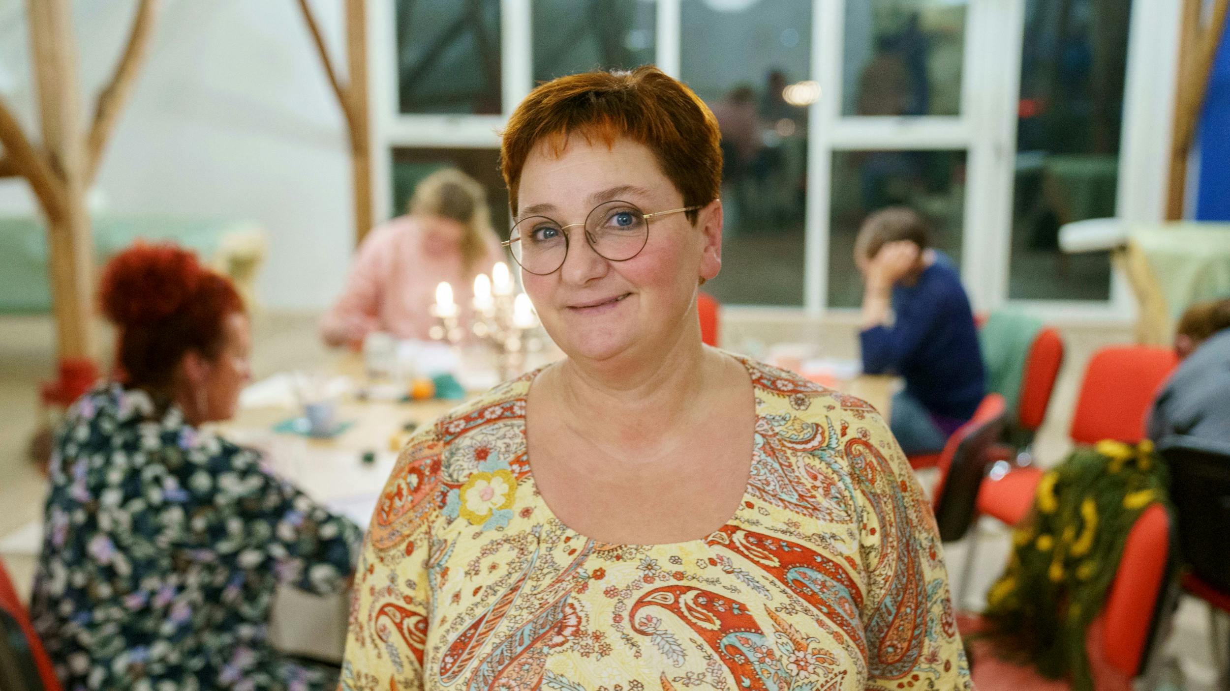 Kvinde med kort, rødt hår smiler i rum med andre kvinder i baggrunden